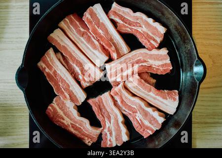 Blick von oben auf den rohen Schweinebauch mit Schichten von Fett und Fleisch in einer schwarzen gusseisernen Pfanne, bereit zum Kochen auf einem Küchenherd. Stockfoto