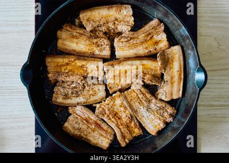 Gewürzte Schweinebauchscheiben, die in einer schwarzen gusseisernen Pfanne gebraten werden, Blick von oben auf fettes Fleisch mit Gewürzen auf einem Küchenherd. Stockfoto