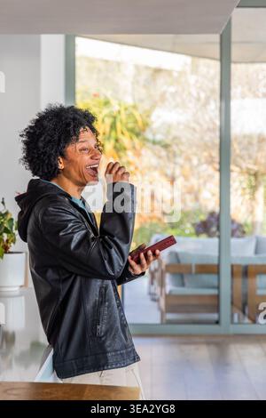 Afroamerikanischer Mann, der sich in Lederjacke an der Haustheke lehnt, lacht mit rotem Telefon, Kopierraum Stockfoto