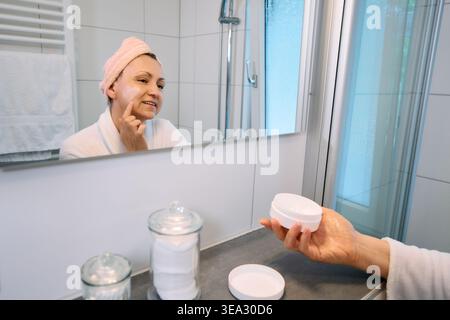 Frau mittleren Alters, die Gesichtscreme aufträgt, in den Spiegel eines hellen Badezimmers blickt, ein weißes Kosmetikglas hält, Hautpflege- und Alterungskonzept. Stockfoto