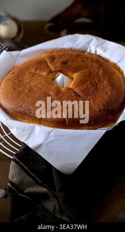 Ein frisch gebackener Ringkuchen liegt in weißem Pergamentpapier auf einem Kühlregal aus Draht. Die weiche, goldene Kruste und die warme Beleuchtung sorgen für ein gemütliches, rustikales Ambiente Stockfoto