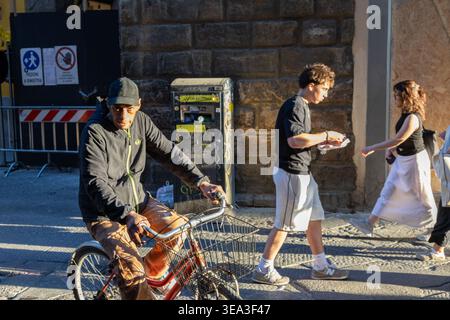 Mann mit Fahrrad und Leute, die auf sonnigem Stadtpflaster mit Steinmauer und Baubarriere im Hintergrund laufen, Florenz Italien. Stockfoto