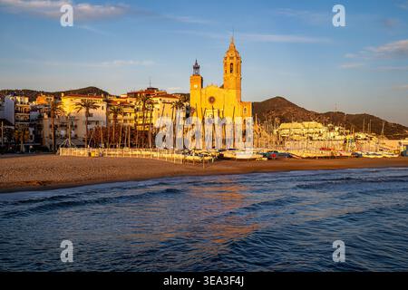 Panoramablick auf Sitges Kirche mit Blick auf Strand und Hafen bei Sonnenuntergang. Warmes goldenes Licht beleuchtet die historische Architektur und die Segelboote auf dem See Stockfoto