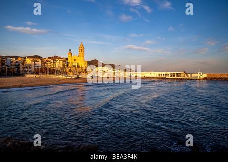 Atemberaubender Blick auf die Sitges Kirche mit Blick auf Fragata Strand bei Sonnenuntergang. Warmes goldenes Licht beleuchtet die barocke Architektur und Segelboote auf dem Sand, Stockfoto
