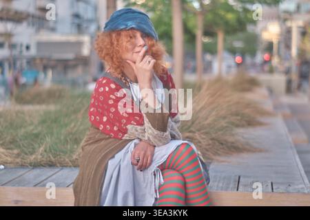 Ein Porträt einer Ingwerfrau mittleren Alters mit lockigen Haaren, die ein exzentrisches Outfit mit gestreiften Strumpfhosen und einer blauen Baskenmütze trägt. Sie sitzt auf einem Holz Stockfoto