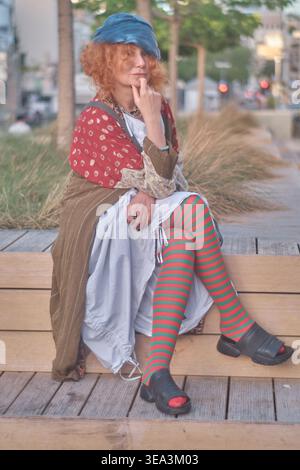 Ein Porträt einer Ingwerfrau mittleren Alters mit lockigen Haaren, die ein exzentrisches Outfit mit gestreiften Strumpfhosen und einer blauen Baskenmütze trägt. Sie sitzt auf einem Holz Stockfoto
