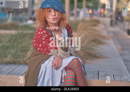 Ein Porträt einer Ingwerfrau mittleren Alters mit lockigen Haaren, die ein exzentrisches Outfit mit gestreiften Strumpfhosen und einer blauen Baskenmütze trägt. Sie sitzt auf einem Holz Stockfoto