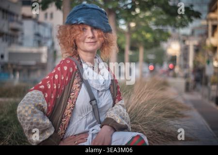 Ein Porträt einer Ingwerfrau mittleren Alters mit lockigen Haaren, die ein exzentrisches Outfit mit gestreiften Strumpfhosen und einer blauen Baskenmütze trägt. Sie sitzt auf einem Holz Stockfoto