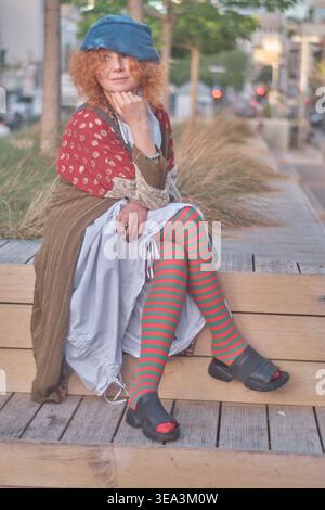 Ein Porträt einer Ingwerfrau mittleren Alters mit lockigen Haaren, die ein exzentrisches Outfit mit gestreiften Strumpfhosen und einer blauen Baskenmütze trägt. Sie sitzt auf einem Holz Stockfoto