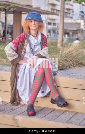 Ein Porträt einer Ingwerfrau mittleren Alters mit lockigen Haaren, die ein exzentrisches Outfit mit gestreiften Strumpfhosen und einer blauen Baskenmütze trägt. Sie sitzt auf einem Holz Stockfoto