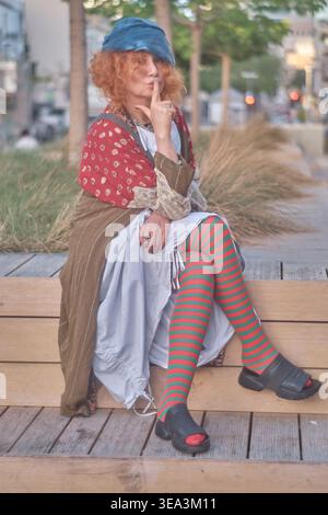 Ein Porträt einer Ingwerfrau mittleren Alters mit lockigen Haaren, die ein exzentrisches Outfit mit gestreiften Strumpfhosen und einer blauen Baskenmütze trägt. Sie sitzt auf einem Holz Stockfoto