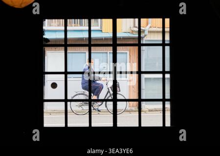 Durch einen dunklen Gitterrahmen fährt ein älterer Mann auf einem Fahrrad eine ruhige Straße in Japan hinunter. Die Silhouette erzeugt eine starke Frame-in-Frame-Ansicht. Stockfoto