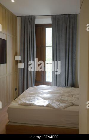 Ungebautes Doppelbett in einem modernen Boutique-Hotelzimmer mit gemusterten grauen Vorhängen und natürlichem Licht, das durch eine Holzbalkontür, Granada, fließt Stockfoto