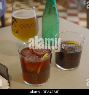 Zwei Eisvermut-Drinks mit Zitronenscheiben, ein Glas El Águila-Bier und eine grüne Flasche Mineralwasser auf einem Café-Tisch in Granada, Andalusien, Spanien. Stockfoto