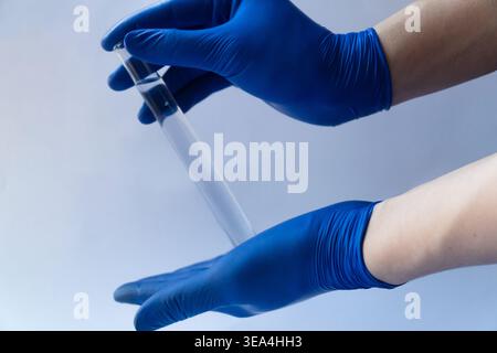 Zwei Hände tragen blaue Nitrilhandschuhe und halten vorsichtig ein mit klarer Flüssigkeit gefülltes Glasröhrchen. Stockfoto