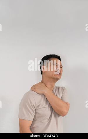 Ein asiatischer Mann mittleren Alters zuckt, während er seine Schulter umklammert, was Nackenverspannungen, Muskelverspannungen, Steifheit und Oberkörperbeschwerden gegen einen cl veranschaulicht Stockfoto