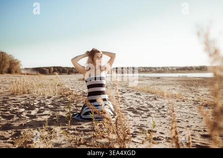 Weiße Frau mit langen blonden Haaren und hinter dem Kopf erhobenen Händen sitzt am Sandstrand, trägt trägerloses schwarz-weiß gestreiftes Kleid und sieht aus Stockfoto