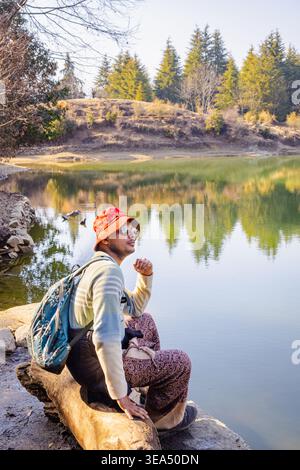 Eine Frau mit Hut und Rucksack sitzt auf einem Baumstamm am See und genießt die friedliche Umgebung des Tungnath-Tempels in Uttarakhand, Indien. Stockfoto