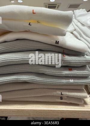 Gestapelte graue und weiße gefaltete Pullover mit Größenetiketten auf dem Hintergrund des Displayregals. Mobiles Foto Stockfoto