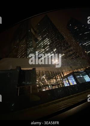 Moderne Wolkenkratzer bei Nacht mit beleuchteten Fenstern und Regentropfen auf Autoglas Stockfoto