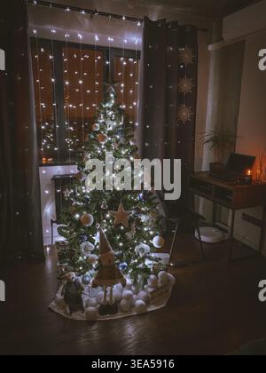 Gemütlicher Weihnachtsbaum, dekoriert mit Lichtern und Ornamenten in einem warmen Wohnraum bei Nacht, festliche Feiertagsatmosphäre. Stockfoto