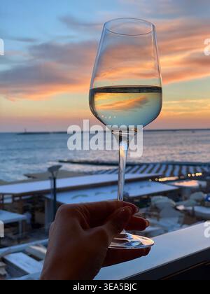 Hand hält ein Glas Weißwein vor einem wunderschönen Sonnenuntergang am Meer, mit weichem pastellfarbenem Himmel im Glas und ruhigem Ozean im Hintergrund Stockfoto