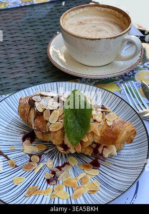 Frisches Croissant mit Mandelflocken und Sahnefüllung, serviert mit einer Tasse Kaffee auf einem Café-Tisch im Freien, für eine gemütliche Frühstücks- oder Brunch-Atmosphäre Stockfoto