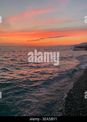 Farbenfroher Sonnenuntergang über dem Meer mit Wellen und Menschen, die in der Nähe von Kieselstrand schwimmen Stockfoto