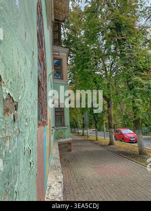 Altes Wohngebäude mit abblätternder Lackfassade und Bürgersteig mit Bäumen und geparktem Auto in der Stadtstraße Stockfoto