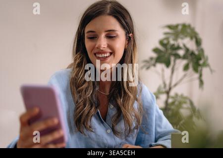 Lächelnde junge Frau, die zu Hause ihr Smartphone benutzt, in einer hellen, ungezwungenen Innenumgebung. Mobile Kommunikation, soziale Medien, Messaging und modernes digitales Leben Stockfoto
