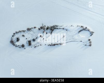 Aus der Vogelperspektive auf eine kleine schneebedeckte Insel mit isolierten Holzhütten und kargen Bäumen, umgeben von einer riesigen gefrorenen Landschaft in Rovaniemi, Lappland, F Stockfoto