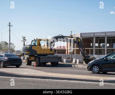 Gelber Radbagger auf einer Straßenbaustelle mit vorbeifahrenden Autos und einem neuen Gebäude im Hintergrund Stockfoto