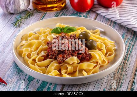 Eine Nahaufnahme hausgemachter Tagliatelle-Pasta mit einer herzhaften Bolognese-Fleischsauce. Garniert mit grünen Basilikumoliven in einer weißen Schüssel auf einem rustikalen Holztisch Stockfoto