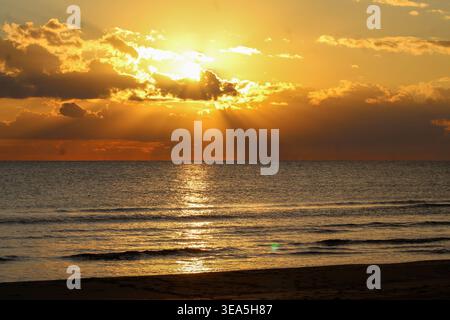 Goldener Sonnenuntergang über dem ruhigen Meer mit dramatischen Wolken und strahlenden Sonnenstrahlen, die durchbrechen, schaffen eine ruhige Küstenlandschaft. Stockfoto