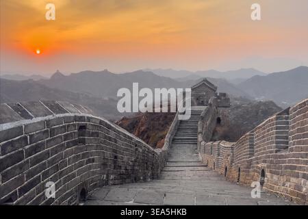 Die Chinesische Mauer bei Sonnenuntergang. Der Sonnenuntergang leuchtet über alten Steinmauern. Treppen führen nach oben durch zerklüftete Berggipfel. Goldenes Licht färbt den Himmel in sanften bernsteinfarbenen Tönen. Weit entfernte Türme stehen stolz inmitten nebeliger Hügel Stockfoto