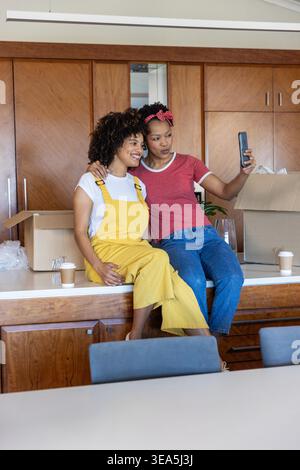 Verschiedene Freundinnen sitzen in der Küche in gelben Overalls und machen Selfie mit Telefon zwischen Boxen. Duo, Smartphone, auspacken, packen, Zuhause, Insel, Arbeitsplatte Stockfoto