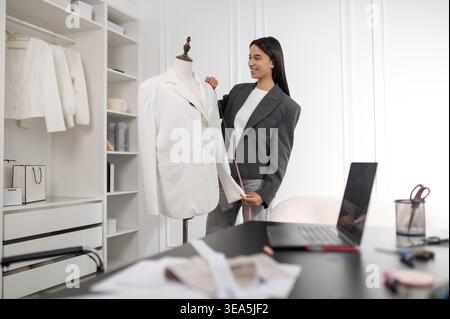 Langhaarige junge Frau mit einer weißen Jacke, die in einem Designstudio neben der Puppe steht Stockfoto