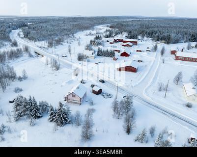 Aus der Vogelperspektive auf traditionelle rote Holzhäuser und eine schneebedeckte Straße, die eine Brücke über einen gefrorenen Fluss in einer winterlichen Waldlandschaft in Lappland, Fi, überquert Stockfoto