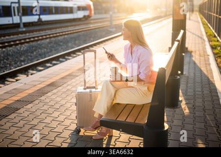 Elegante Geschäftsfrau, die am Bahnhof auf den Zug wartet, SMS per Handy sendet, Kaffee hält, entspannt und lächelt während der Geschäftsreise. Stockfoto