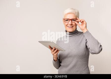 Ältere Frau lächelt, während sie ein Tablet und eine Brille in einem gemütlichen Innenraum benutzt Stockfoto