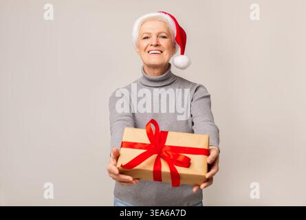 Lächelnde ältere Frau mit Weihnachtsmütze präsentiert ein Weihnachtsgeschenk Stockfoto