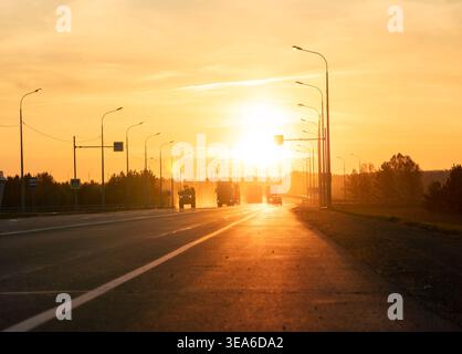 Golden-Hour-Licht beleuchtet Fahrzeuge, die bei Sonnenuntergang auf einer breiten Straße fahren, die von Straßenlaternen und weit entfernten Bäumen gesäumt ist Stockfoto