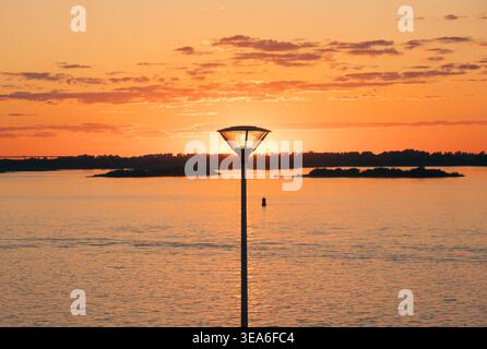 Die Silhouette der Straßenlaterne vor einem leuchtenden orange-gelben Himmel während eines wunderschönen Sonnenuntergangs über einem breiten Fluss Stockfoto