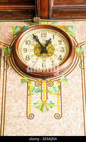 Vintage-Wanduhr mit Holzrahmen an kunstvoll verzierten Mosaikfliesen, Jugendstilgeblümten, warmen Tönen, Inneneinrichtung aus nächster Nähe. Stockfoto