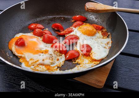 Nahaufnahme von zwei Spiegeleiern, die mit Kirschtomaten in einer Pfanne auf einem dunklen Tisch gekocht wurden, mit einem Holzspachtel. Frühstücksszene Stockfoto