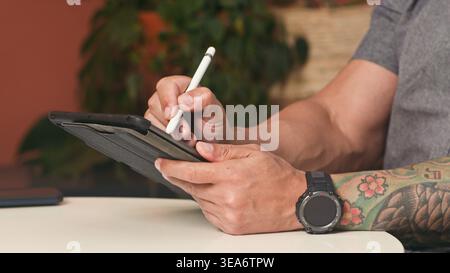 Mann, der ein Tablet mit Stift im zwanglosen Arbeitsbereich mit tätowiertem Arm und Smartwatch verwendet Stockfoto