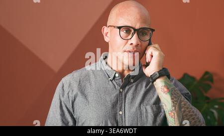 Kahler Geschäftsmann Mit Brille, Der Am Telefon Spricht, Während Er Im Modernen Büro Tätowierte Arme Zeigt Stockfoto
