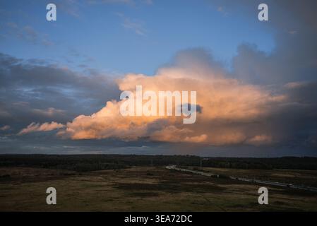 Dramatische Sturmwolken sammeln sich über einer weiten ländlichen Landschaft mit einer gewundenen Straße, die ein Feld und einen dunklen Wald darunter durchquert. Warmes Sonnenlicht, das eine auffällige Wolkenbildung während eines lebhaften Sonnenuntergangs beleuchtet Stockfoto