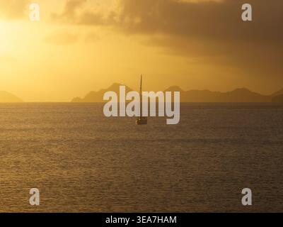 Aus der Vogelperspektive eines einsamen Segelboots auf dem ruhigen Wasser von Anse A La Mouche während eines goldenen Sonnenuntergangs mit Inselsilhouetten am Horizont der Seychellen. Stockfoto