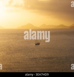 Aus der Vogelperspektive eines einsamen Segelbootes auf dem ruhigen Meer von Anse A La Mouche während eines goldenen Sonnenuntergangs mit Bergsilhouetten im Hintergrund Seychel Stockfoto
